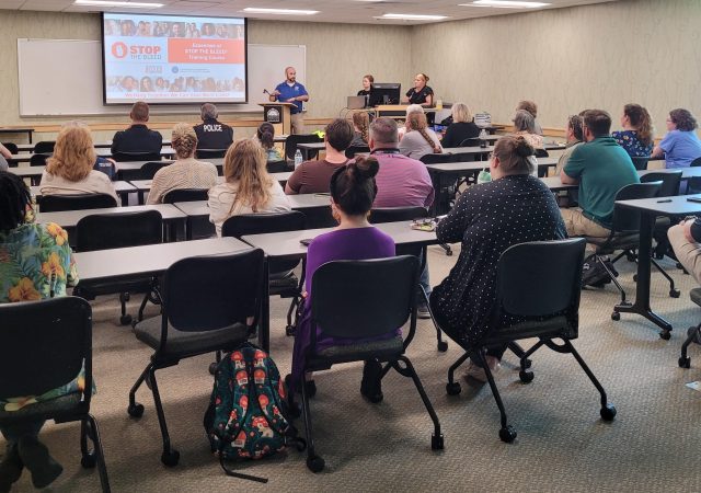 PHOTO - Classroom setting. Three instructors at the front of the room with a presentation on the big screen - "Stop the Bleed" training title; numerous participants sitting in classroom facing instructors.